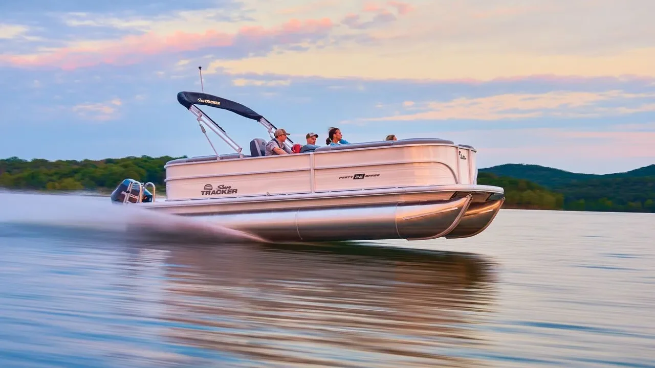 1 (1) A white Sun Tracker Party Barge 22 pontoon boat with multiple passengers, cruising on a calm lake at sunset with a pink and orange sky.