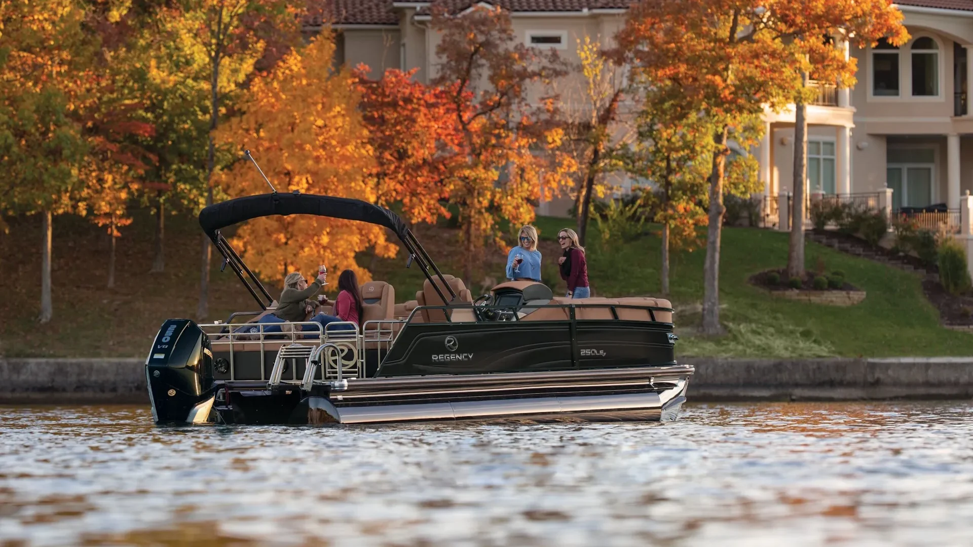 Regency-scaled A luxurious black Regency 250 LE3 pontoon boat with four passengers, idling near a shoreline with large houses and vibrant autumn foliage.